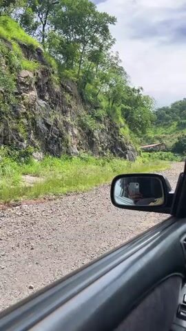 The video shows a view from inside a car looking out the passenger side window as the car drives down a gravel road. To the left of the road is a steep, rocky hillside covered in green vegetation. To the right of the road, further in the distance, are some small buildings on a hillside. The side mirror of the car is visible, reflecting the interior of the car and a person. The sky is overcast with clouds. The car is moving forward on the road.