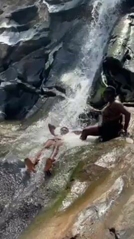 A waterfall is shown, and three people are in the water. One person is lying down while getting hit by the water. Another person is sitting with their legs in the water. The third person is sitting in the water. The background consists of gray rocks and running water.