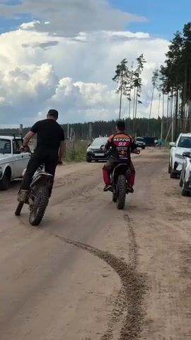 The video shows two individuals riding dirt bikes on a dirt road. The person in the foreground on the right is wearing a black and orange racing suit with the number 53. The person in the background on the left is wearing a black t-shirt and black pants. Both riders are kicking up dust as they move forward. The road is lined with trees and there are several cars parked on the side of the road. The sky is blue with some white clouds. The video appears to be taken from behind the riders.