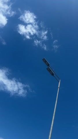 The video shows a street light against a bright blue sky dotted with sparse white clouds. The camera zooms in on the two rectangular LED lights atop a metal pole. The lights are off. The camera focuses on the grid of LEDs within the rectangular housing, framed by a toothed edge.
