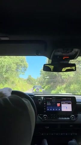 The video is filmed from the interior of a vehicle, showing the perspective of a driver. The driver's hand is visible on the steering wheel. Ahead, another vehicle, a light-colored SUV, is visible on a rural road surrounded by trees and foliage. The sky is blue and clear. The vehicle's dashboard displays a touchscreen infotainment system.
