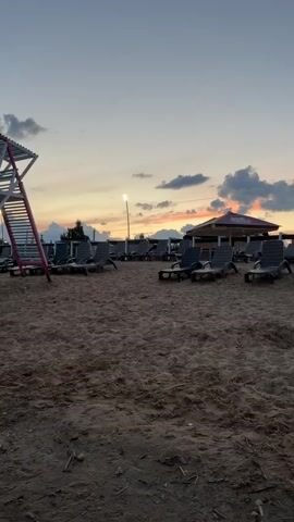 This is a vertical, outdoor, ground-level video of a sandy beach at dusk. The sky is a gradient of pale orange, pink, and blue, with scattered clouds. In the foreground, the sand is slightly textured, showing indentations and subtle variations in color. Further back, rows of empty lounge chairs are arranged on the sand. On the left, a tall, white lifeguard stand with red accents is partially visible. In the background, a few low buildings with awnings and a single tall lamppost with a bright light are visible, along with some trees. The overall impression is a quiet, empty beach as evening sets in.