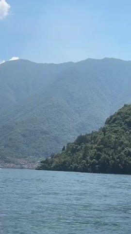 The video shows a scenic view of a large body of water with mountains in the background. The water is a deep blue and appears calm with gentle ripples. The mountains are covered in lush green vegetation, indicating a temperate climate. In the distance, on the left side of the frame, a small town or village with red-tiled roofs can be seen nestled along the shoreline. The sky is a clear blue with a few wispy clouds. The perspective suggests the camera is positioned on a boat or near the water's edge, looking out towards the landscape. The lighting indicates it is daytime, likely late morning or early afternoon, with the sun casting a bright glow on the scene.