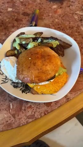 The video shows a plate of food on a reddish-brown marble table.  The plate has a burger with cheese, sauce, and a bun.  There are also potato wedges with a green sauce, and a dollop of white sauce. There is a vape pen near the top left of the plate. The camera zooms in on the burger.