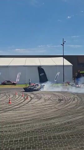 A black drift car with red and yellow accents is performing a donut maneuver in a paved outdoor area. The car is surrounded by orange traffic cones arranged in a circle, and it is producing a large amount of smoke from its tires. In the background, there is a large white tent structure with several flags displaying logos. Two other cars are parked in the distance to the left of the tent. The sky is blue with some scattered clouds.