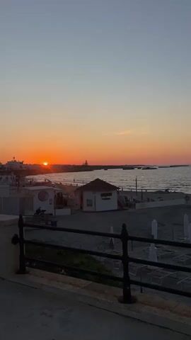 This is a vertical video of a sunset over a beach. The sun is a bright orange orb low on the horizon, casting a warm glow over the scene. The sky is a gradient of orange, pink, and blue. In the foreground, there is a dark railing and a walkway. Beyond the railing is a sandy beach with lounge chairs and umbrellas. There are a few small buildings on the beach, one of which appears to be a concession stand with signage. The ocean stretches out to the horizon, with small waves lapping at the shore. A few boats are visible in the distance. The overall impression is one of a peaceful and serene evening at the beach.