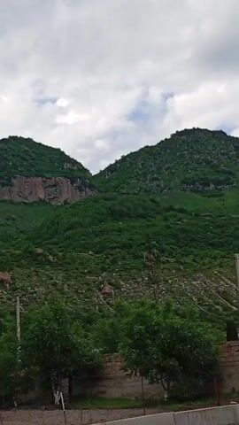 The video presents a panoramic view of a lush, green mountain range under a partly cloudy sky in Sayukovo, Russia. The dominant feature is a large mountain or hill, densely covered with varied vegetation, including shrubs, bushes, and scattered trees, with visible rocky outcrops and cliff faces. Near the summit of the main peak, two distinct communication towers or similar structures are discernible against the sky.

The sky is predominantly overcast with light gray and white clouds, although patches of brighter sky can be seen through breaks in the cloud cover, suggesting daytime.

In the foreground, a glimpse of a built environment is visible. To the left, a small, leafy green tree stands before a light-colored concrete block wall. Further to the right, a tall, white utility pole extends upwards, positioned in front of a low, light-brown brick wall that stretches across the lower part of the frame. Behind this wall, several dark green, conical evergreen trees are planted, and a red-tiled roof of a building is partially visible.

The camera executes a slow, deliberate pan from left to right, smoothly revealing more of the mountain's expanse and the residential structures nestled at its base. No people or visible text appear in the video. The scene captures a tranquil, natural landscape interspersed with subtle signs of human presence.
