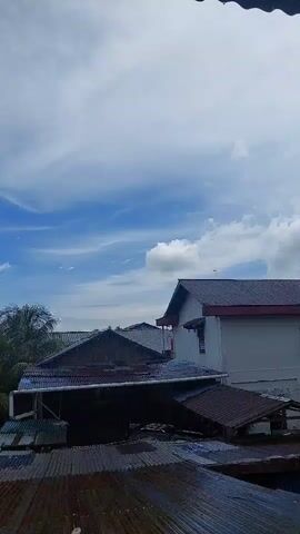 A time-lapse video shows a cloudy sky with a blue sky visible. There are several houses with corrugated metal roofs. The roofs are wet, indicating it may be raining or has recently rained. There are some trees visible in the background.