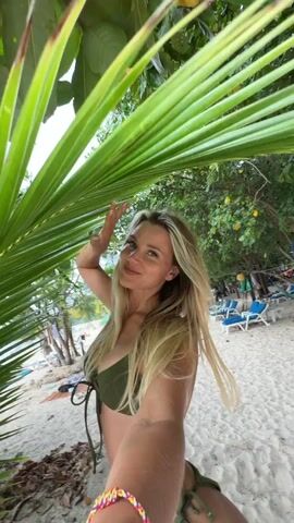 A blonde woman wearing a green bikini is taking a selfie on a beach. She is partially obscured by a large palm frond in the foreground. Behind her, the beach is visible with lounge chairs and umbrellas. The woman is smiling and appears to be gesturing with her hand. The lighting suggests it is daytime, possibly overcast.