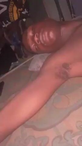 A shirtless Black man lies on a bed. His eyes are closed. An arm is extended to the side, revealing armpit hair. A cell phone and luggage are visible on the bed nearby. The room is dimly lit.