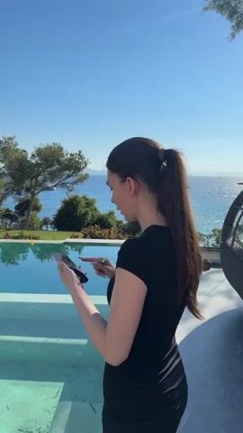 A woman in a black dress stands by an infinity pool, looking at her phone. The pool is filled with clear, turquoise water and has steps leading into it. Beyond the pool, there is a view of the ocean, with trees and foliage surrounding the area. The sky is clear and blue, indicating a sunny day.