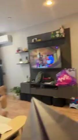 The video begins with a close-up of a brown paper object, possibly a bag or large envelope, held in front of the camera. The camera is held inside a room, and the image quickly pans up and to the left to show a child throwing a paper airplane. The child, a young boy with blond hair, stands on a bed and is wearing a diaper. He is holding a small yellow toy car. He smiles at the camera. The bed has a light tan or beige surface with black and white zebra-striped pillows. There are also paper airplanes on the bed. A window is visible in the background. The window has sheer white curtains, and outside the window, bare trees are visible. The room has hardwood flooring, and a television is visible on the wall.