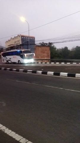 This is a vertical video taken from a slightly elevated perspective, likely from a vehicle or overpass, looking down and to the side at a street scene. The time of day appears to be dusk or early dawn, with overcast skies and a single street light illuminated on the left side of the frame.

In the middle ground, on the left side of the street, a white bus is parked. The bus has blue tinted windows and its headlights are illuminated, casting a bright glow. There are markings on the side of the bus, including red lettering that reads "MAJU". Adjacent to the bus, there are several buildings. One building in the background is made of brick and appears to be older, possibly under construction or in disrepair, with exposed brickwork and an unfinished look. To the right of this brick building is a lighter colored building, which looks more modern with windows.

The street itself is paved with asphalt and features a distinct white line separating lanes and a black and white striped curb along the edge. The perspective creates a sense of depth, with the street stretching into the distance. Power lines are visible running diagonally across the upper left portion of the frame. The overall environment suggests an urban or suburban setting. No people are clearly visible in the frame. The bus appears to be stationary.