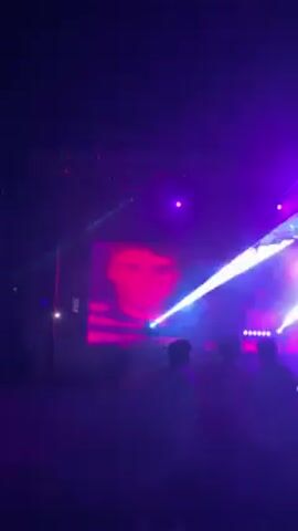 The video shows a night scene, likely a party or club setting, with people dancing and socializing under colorful lighting. Initially, the camera faces a stage with a spotlight and silhouettes of figures. A purple-blue laser light beam scans across the scene. As the camera pans up and away, the location appears to be outdoors, with red fabric draped above the crowd. The crowd is diverse, with people dressed in both white and colored clothing. Some are standing in groups, conversing, while others are moving to the music. The atmosphere is lively and energetic, amplified by the lighting and music.
