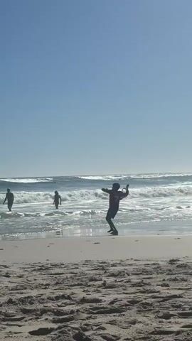The video shows a beach scene on a sunny day. In the foreground, a person is standing on the sand, facing the ocean, and appears to be throwing an object into the air. In the background, two people are walking into the ocean, one carrying a surfboard. Waves are visible in the ocean. The sky is clear and blue.