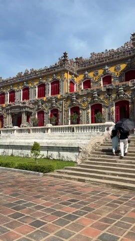 The video shows a young woman in a black tank top and red skirt spinning in front of a large, ornate yellow building with red windows. She is wearing a black baseball cap, sunglasses, and white sneakers with white socks. She has a red bag slung across her chest. She is spinning in a patterned courtyard paved with red and brown square tiles. To her right, a set of stone stairs leads up to the building, and several people are walking up the stairs, one of whom is holding a black umbrella. The building is elaborate with decorative carvings and a yellow facade. The sky is blue with some clouds.