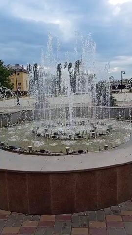 The video shows a fountain in operation. The fountain has multiple jets spraying water upwards and outwards, creating a curtain of water. The fountain is circular and appears to be made of concrete or stone. There are lights around the base of the fountain, some of which are illuminated with colored light. In the background, there are buildings and trees. The sky is cloudy. The video is shot from a fixed perspective.