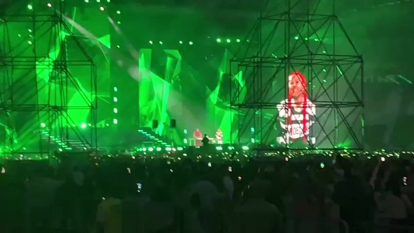 The video shows a concert with a large audience in the foreground. The stage is lit with vibrant green lights and features large screens displaying abstract geometric shapes and a performer. The performer on the screen is a woman with red hair wearing a white outfit with black detailing. There are also smaller figures on the stage in the distance, likely other performers or crew members. The stage structure is elaborate, with black scaffolding-like elements framing the screens and the performer. Many audience members are holding up their phones, possibly recording the event. The overall atmosphere appears to be energetic and exciting.