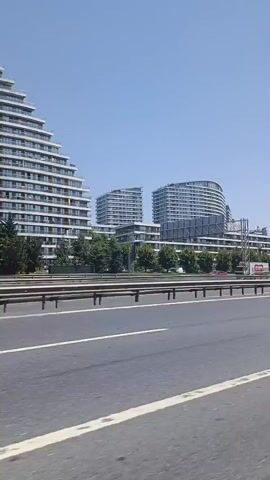 The video shows a daytime scene from a moving vehicle. In the foreground, a gray asphalt road with white lane markings is visible. A metal guardrail separates the road from the background. The background features a cluster of modern, multi-story buildings with white facades and glass balconies. Trees and greenery are interspersed between the buildings. The sky is clear and blue. Several vehicles, including cars and a truck, are driving on the road. The overall impression is of an urban environment with contemporary architecture and a highway.
