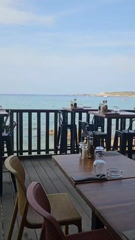 A vertical, slow-panning video shows an outdoor restaurant seating area with a view of the ocean. The tables are made of wood and are set with glasses, cutlery, salt and pepper shakers, and bottles of oil and vinegar. There are various chairs around the tables, including some made of rattan and plastic. In the background, there is a wooden railing overlooking the sea, with several boats visible in the distance. The sky is a clear blue, and the water appears calm.