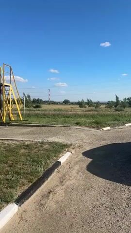 A vertical video opens on a wide, clear blue sky with scattered white clouds. Below the sky, a landscape unfolds with dry, brown grass and sparse green vegetation. In the distance, a tall, red and white smokestack stands against the horizon. To the left of the frame, a bright yellow metal structure with a railing rises vertically, casting a shadow on the ground. The foreground features a gravel path with a black and white curb. As the video progresses, the camera pans to the right, revealing a line of cars parked in a grassy area with trees. The cars are mostly white SUVs and black sedans. The background shows a body of water and more trees, with a few buildings visible further in the distance. The overall scene depicts an outdoor environment on a sunny day, likely in a rural or industrial setting.