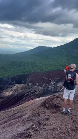 A person stands at the edge of a volcanic crater, looking out over a vast, lush green landscape under a cloudy sky. The person is wearing a black tank top with white text that reads "HIKE SIDE TRIP" and white shorts. They are holding a bright orange bottle. The crater itself is a deep, reddish-brown and appears to be made of volcanic rock and ash. The surrounding hills are covered in dense green vegetation, and in the distance, rolling hills and a hazy horizon are visible. The sky is filled with dramatic, dark clouds, suggesting a storm might be approaching. The overall impression is one of awe and rugged natural beauty. The time of day appears to be late afternoon or early evening, given the lighting and cloud cover. The location is Rota, Nicaragua, a place known for its volcanic features. The scene evokes a sense of adventure and exploration.