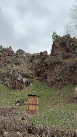The video shows a rugged, rocky hillside on a cloudy day. The hillside is covered in patches of green grass and dry brush. At the base of a cliff is a cave-like structure with a wooden door or barrier. A metal chimney stands in the foreground. There are no people visible in the frame. The camera zooms slightly closer to the cave.