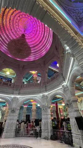The video shows the interior of what appears to be a temple or large religious building. The walls and pillars are white with intricate carvings. People can be seen milling around on the ground level and on a balcony that runs along the second floor. The most striking feature is the domed ceiling which is elaborately decorated with concentric circles lit in alternating colors: pink, orange, yellow, and green. A large chandelier hangs from the center of the dome. There are fans mounted on the walls. The lighting is a combination of natural light and colored LED lights that accent the architectural details. The overall impression is of a place of worship that is both grand and modern.