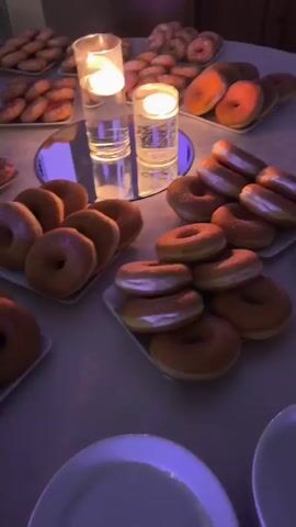 The video displays a dessert buffet table laid out with numerous donuts arranged on white platters. The lighting is dim and purple-toned. Two glass candle holders with lit white candles are placed on the table. In the background, another buffet table with more food items and a person in a uniform can be seen. The overall environment appears to be an indoor event setting.