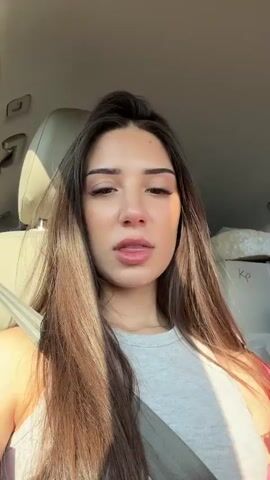 A young woman with long, dark hair is seated in a vehicle, wearing a light gray tank top and a seatbelt. She is holding a plastic cup with a dome lid and a black straw, containing a light brown beverage. She looks at the camera and speaks while holding the cup, at one point extending her tongue. The car's interior is visible, with sunlight coming through the windows.
