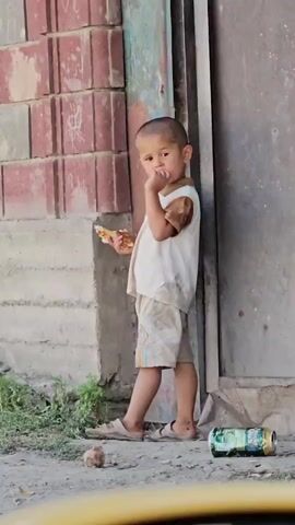 A child stands leaning against a building. The child is holding a piece of food in their left hand and has their right hand near their face. The child is wearing a white sleeveless shirt with brown accents on the shoulders and light-colored shorts. The child is barefoot, wearing sandals. A discarded aluminum can is on the ground to the right of the child. There is some greenery and dirt on the ground. The building has a brick wall on the left and a metal door on the right.