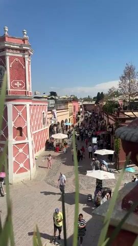 The video shows a busy pedestrian street in what appears to be a historic town or city, possibly in Mexico or South America. The street is paved with cobblestones and lined with buildings of various colors, mostly in shades of pink, yellow, and red. Many people are walking along the street, some are shopping, others appear to be tourists. Several white umbrellas provide shade for vendors or small businesses. A tall, red-and-white patterned building stands at the edge of the frame. The sky is blue and clear. In the foreground, out-of-focus green plants partially obscure the view.