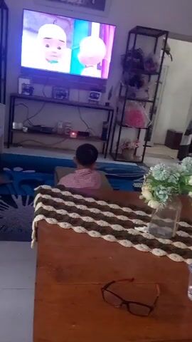 A boy sits on the floor facing a large television screen which is displaying an animated program. He is wearing a pink shirt. In the foreground, a wooden coffee table is visible with a knitted runner and a vase of flowers. A pair of glasses rests on the coffee table. The room appears to be a living room.