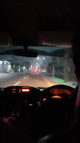The video provides a first-person perspective from inside a car at night, looking forward through the windshield. The dashboard is visible in the foreground, with orange illuminated gauges and a red display. The steering wheel, also illuminated with orange, is partially visible. The driver's back and shoulder are in the lower right corner of the frame, suggesting the camera is positioned behind or beside them. Outside the car, a road is visible with streetlights and the lights of other vehicles, indicating urban or suburban traffic. The scene is dark, with artificial lights creating reflections on the windshield and illuminating the road ahead.