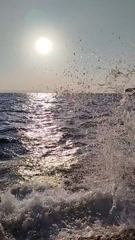 The video shows a body of water, likely the ocean, with waves crashing against what appears to be a shore. The sun is visible in the upper left corner, casting a bright reflection on the water. The overall scene is one of natural beauty, possibly captured from a boat or a cliff overlooking the water. There are no people or specific objects clearly identifiable in the footage, focusing solely on the dynamic interaction of the water and light.