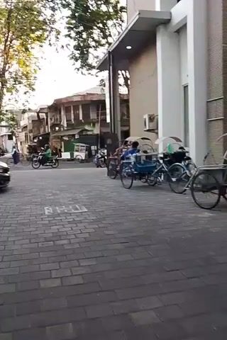 The video shows a street scene with a dark-colored hatchback car turning in the foreground. On the right side of the street, there are several bicycle rickshaws parked, some with passengers. In the background, there are buildings and trees. People can be seen riding motorcycles and walking. The street is paved with cobblestones. The lighting suggests it is daytime.