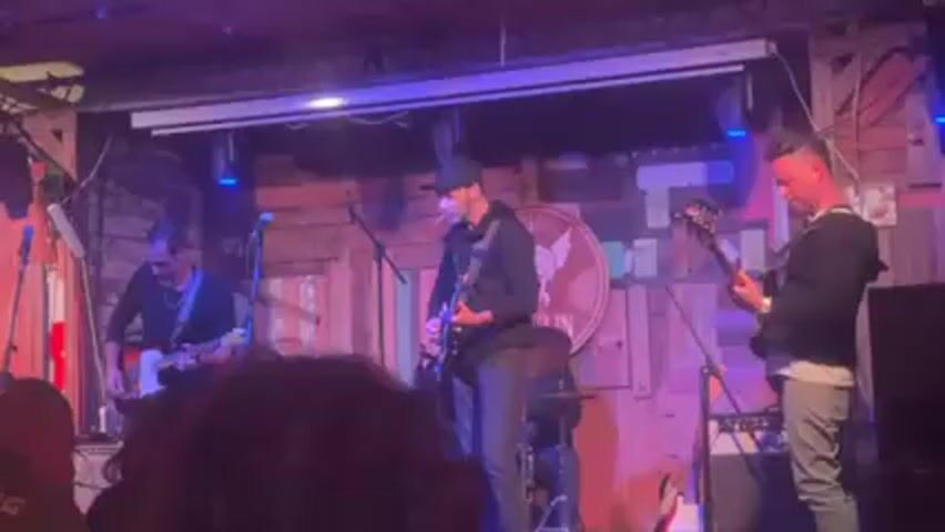 The video shows a live band performing on a small stage. Three men are visible: one on the left playing an electric guitar, another in the center playing another electric guitar and wearing a black baseball cap, and a third on the right playing a bass guitar. The stage backdrop is a wooden wall with a circular logo featuring an eagle. Stage lighting is predominantly blue and pink. A few microphones are set up. In the foreground, the back of someone's head is visible. The room appears to be a bar or small music venue.