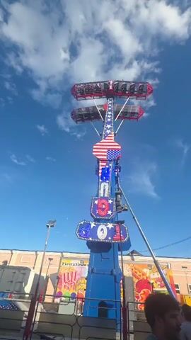 A wide shot captures a tall, blue amusement park ride with an American flag design at its center. Two gondolas, each carrying passengers, are attached to the ends of a horizontal arm, which is suspended from a vertical central tower. The ride is in motion, with the horizontal arm rotating and swinging the gondolas. The sky above is blue with scattered white clouds. In the background, there are buildings and other carnival attractions, including a sign that reads "Fiesta Shows." Several people are visible at the base of the ride, some standing and observing, others appearing to be waiting in line. The overall scene is brightly lit, suggesting it is daytime.