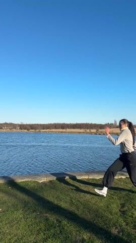 A young woman is practicing a martial arts kick near a body of water on a sunny day. She is wearing sunglasses, a gray sweatshirt, black pants, and white sneakers with black accents. Her hair is tied back in a ponytail. She stands on a grassy area next to a concrete edge bordering a blue lake or river, against a clear blue sky. In the background, there are some trees and brush along the distant shore.