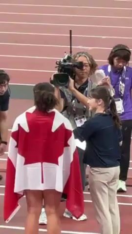 On a red track, a woman draped with a Canadian flag stands facing a young woman in a dark blue shirt and khaki pants. The young woman appears to be conversing with the athlete.  A man behind the athlete holds a professional camera and seems to be filming. There are other people around, one squatting and another standing in a purple shirt and badge. The camera operator wears glasses, and some of his hair is styled in short dreadlocks.  The setting appears to be a stadium or sports venue, judging by the running track markings.