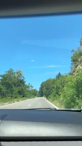 The video shows a view from inside a car as it travels along a paved road. The sky is clear and blue with a few wispy clouds. Dense green trees line both sides of the road. The road appears to be in a rural or natural setting. The camera is positioned behind the dashboard of the car, looking forward through the windshield. The video is short, lasting only a few seconds, and appears to capture a moment of the car's journey.