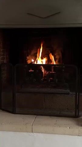 The video shows a fireplace with a fire burning behind a black metal screen. The fireplace appears to be brick, and the hearth is made of light-colored stone tiles. Flames are visible above the grate inside the fireplace. The fire is flickering and creates a warm, orange glow. The surrounding environment is dimly lit. In the background, muffled voices can be heard.