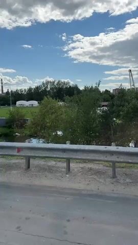 The video shows a rural landscape with a blue sky and clouds. In the foreground, a metal guardrail runs along a paved surface. Beyond the rail, there is a body of water with some white objects floating on it. Further in the background, there is a green field with several white tents. A small red and white aircraft is visible on the grass. Trees and some buildings are scattered throughout the scene. The video appears to be shot from a moving vehicle, as indicated by the slight blur in some parts of the footage.
