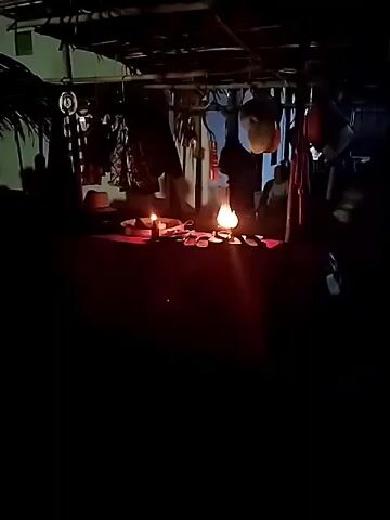 A dimly lit scene, likely outdoors under a canopy. A red surface, possibly a table or stall, is illuminated by a kerosene lamp in the center. Several items are arranged on the red surface, including a candle to the left of the lamp. The background shows hanging items, possibly clothing or merchandise, and palm fronds to the left. Silhouettes of people are visible in the foreground and background, with one person in the mid-ground appearing to interact with the items on the red surface, possibly performing a ritual. The audio indicates someone speaking, with the phrase "dua kali putaran" (two rotations) repeated twice, followed by "balik lagi satu kali" (again one time).