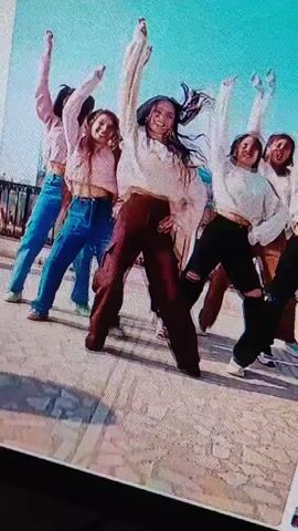 The video displays a group of seven young women dancing in front of a bright, hazy sky and a light-colored, possibly paved, outdoor surface. They appear to be performing a choreographed dance routine. The woman in the foreground, in the center, wears a white top and burgundy pants, her body angled to the right with her right leg extended. To her left, a woman in blue pants and a light-colored top faces forward, her arms raised. Behind the central dancer, several other women in similar light-colored tops and various colored pants are visible, some with their arms raised or bent. The overall mood is energetic and youthful, with the dancers' expressions suggesting enjoyment. The image quality is somewhat grainy, indicating it may be a low-resolution recording.