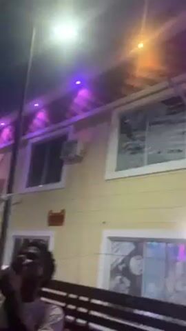 The video shows an outdoor scene at night. A person in a white shirt is holding a phone up high, appearing to take a photo or video of a building. The building is painted light yellow and has several windows and air conditioning units. There are purple lights above the windows. Next to the person filming, another person in a striped shirt and cap is looking at their phone. A white car is parked near the building. The overall lighting is dim, with streetlights and building lights illuminating the scene.