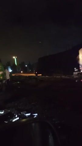 The video is a dark, nighttime scene. The primary focus is on a large, tarp-covered truck parked on what appears to be a dirt or gravel lot. The truck is illuminated by its own taillights and some ambient light from the surroundings, including a bright green light visible in the distance. Another truck, darker and less distinct, is partially visible on the right side of the frame. There are puddles of water on the ground, reflecting the available light. In the foreground, out of focus, is a portion of a vehicle, possibly the interior of a car looking out. There are other vehicles and structures faintly visible in the background, suggesting this is some kind of roadside area or lot. A person is standing near the front of the tarp-covered truck, facing away from the camera. The overall impression is one of a quiet, possibly isolated location at night.