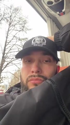 A man is inside what appears to be a bus, leaning his head against his hand and the seat. He is wearing a black jacket and a black baseball cap featuring a white skull design and the word "THRUST". The tree without leaves is visible through the bus window. The bus ceiling contains air conditioning vents. The man looks directly at the camera, his facial expression neutral. His face is closely cropped, with a slight beard.