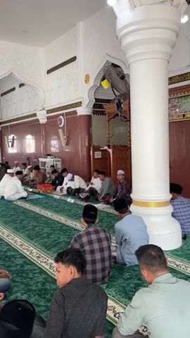 The image depicts a religious gathering of men inside a mosque, likely Mesjid Reubee in Indonesia, during the daytime. The mosque features white walls adorned with intricate decorative relief patterns and large, fluted white columns. The floor is covered with a vibrant green prayer carpet, intricately patterned with cream-colored borders and designs.

In the foreground, several men are seated on the carpet with their backs to the camera. They are dressed in casual attire, including a plaid shirt, a grey long-sleeved shirt, a white t-shirt, and a dark t-shirt, with some wearing peci (prayer caps). They are facing towards the center of the room where a larger group is engaged in an activity.

In the midground, a group of men is seated facing each other. An older man, dressed in a white robe and white peci, sits on a slightly elevated mat or cushion, appearing to lead the gathering. He is facing a group of other men, many of whom are also wearing peci and traditional Indonesian clothing, some in white robes. A small, dark wooden table or chest is placed between these two groups, possibly holding religious texts or other items. More individuals are seated further back along the walls.

In the background on the right side of the frame, a wooden mihrab or minbar (pulpit) structure is visible, decorated with carved patterns. Above it, an Arabic calligraphy inscription from the Quran (Surah Al-Ahzab, 33:56) is prominently displayed: "إِنَّ اللَّهَ وَمَلَائِكَتَهُ يُصَلُّونَ عَلَى النَّبِيِّ ۚ يَا أَيُّهَا الَّذِينَ آمَنُوا صَلُّوا عَلَيْهِ وَسَلِّمُوا تَسْلِيمًا" (Indeed, Allah and His angels send blessings upon the Prophet. O you who have believed, ask [Allah to confer] blessing upon him and greet [him with] a [becoming] salutation). Ceiling fans are mounted on some columns and walls for ventilation. The atmosphere appears calm and attentive, suggesting a religious lesson, discussion, or ceremony.