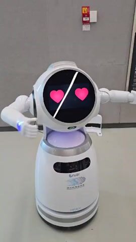 Here's a plain text description of the video:

A white, cylindrical service robot with a circular screen for a face stands on a gray tiled floor against a gray wall. The robot has two arms with articulated fingers. The screen displays two pink hearts. The robot's hands are positioned as if it's giving a hug.  A purple light emanates from the base of the robot.  The robot is labeled with "Cruzr" and "UBTECH."  Background elements include a small red fire alarm box on the wall and a black sign. The robot is stationary. Pop music plays in the background.